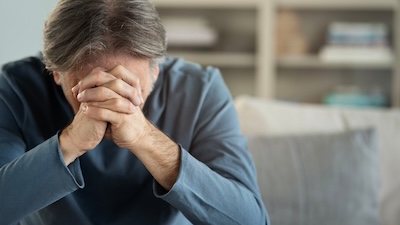 Man over 45 sitting at home with hands covering face appearing overwhelmed by stress or depression